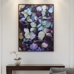 A 60 cm x 90 cm painting of purple, white, and blue hydrangea petals is centrally placed on a white wall in a luxurious reception area. The painting is on a gallery-wrapped canvas and is illuminated by three recessed spotlights above it. Below the painting, a dark wooden console table holds a small white orchid in a textured ceramic pot on the left side. To the right, a gray sofa with matching throw pillows is partially visible, indicating a cozy seating arrangement. The wall features subtle white wainscoting details, enhancing the sophisticated ambiance of the room.