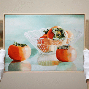 A pair of white-gloved hands holding up a framed painting of persimmons in a glass bowl, with a soft, light blue background. The painting is oriented horizontally and shows two persimmons outside the bowl, one on the left and one on the right, with several more inside the clear, fluted glass bowl. The light source creates reflections on the surface the bowl and fruit are resting on. The hands hold the painting up towards the viewer, with the thumbs positioned on the lower edge of the frame. In the blurry background, there are other framed artworks visible on a wall.