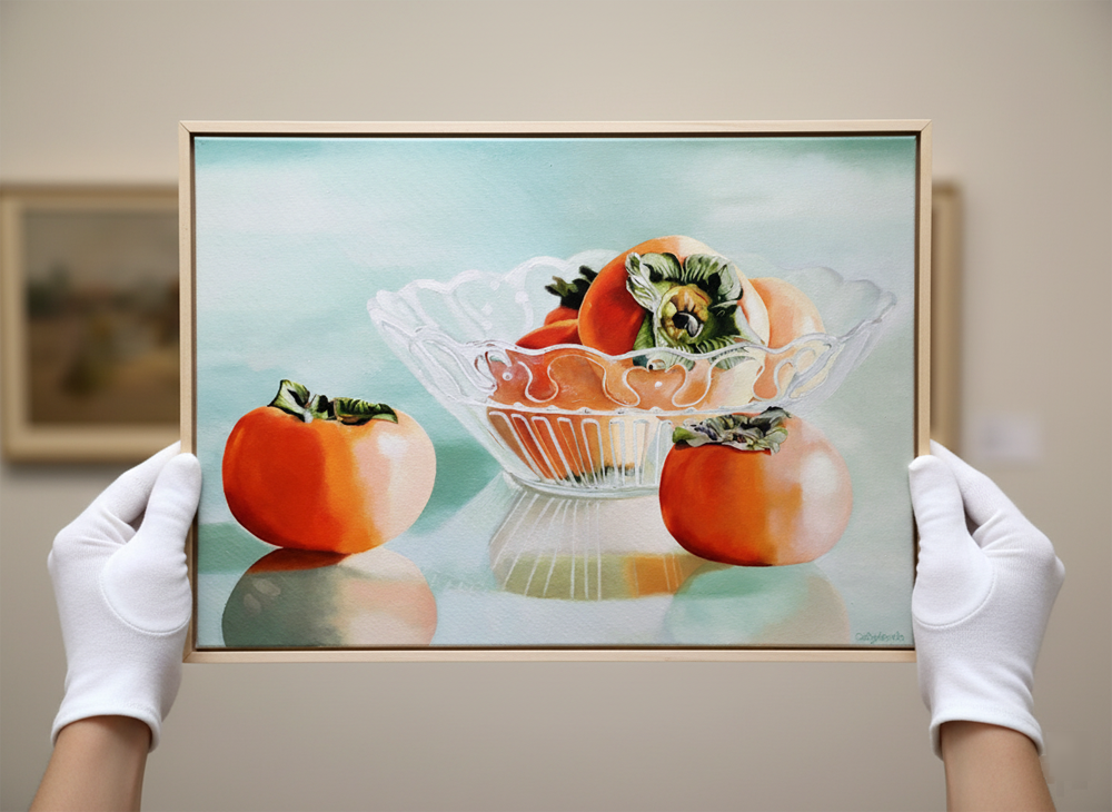 A pair of white-gloved hands holding up a framed painting of persimmons in a glass bowl, with a soft, light blue background. The painting is oriented horizontally and shows two persimmons outside the bowl, one on the left and one on the right, with several more inside the clear, fluted glass bowl. The light source creates reflections on the surface the bowl and fruit are resting on. The hands hold the painting up towards the viewer, with the thumbs positioned on the lower edge of the frame. In the blurry background, there are other framed artworks visible on a wall.
