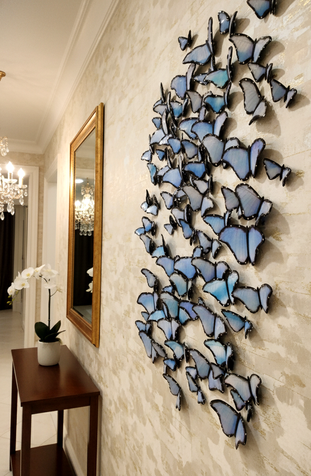 A stunning interior shot shows a large, dynamic wall installation made of numerous blue and white butterflies. The butterflies are arranged in a flowing, upward motion, giving the impression of a fluttering swarm. They vary in size, with some larger pieces and many smaller ones, creating depth and visual interest. The butterflies are affixed to a textured, cream-colored wall that has subtle gold horizontal stripes. To the left of the butterfly installation, a framed rectangular mirror with a gold frame hangs above a dark wooden console table. On the table, a white orchid in a pot adds a touch of natural elegance. In the background, a luxurious chandelier with multiple lights can be seen, hinting at the upscale nature of the space. The lighting in the room is warm, highlighting the details of the butterflies and the elegant decor. The overall impression is one of sophistication and artistic design.