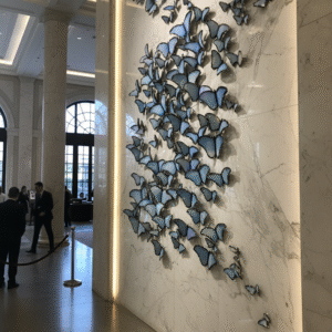 A high-angle, full shot shows a wall covered in a sprawling installation of blue butterflies of various sizes. The butterflies are arranged in a dense, flowing pattern, giving the impression of a swarm in motion. The butterflies themselves are a vibrant blue with hints of purple, and their wings appear to have a slightly metallic or iridescent sheen. Some butterflies are clustered together, while others are more spread out, creating a dynamic visual effect. The background is a plain, light gray surface, which makes the butterflies stand out prominently. The overall impression is one of elegance and delicate beauty.