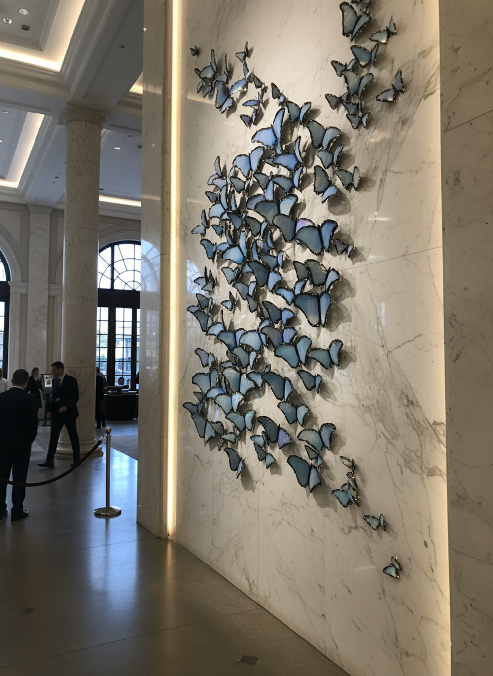 A high-angle, full shot shows a wall covered in a sprawling installation of blue butterflies of various sizes. The butterflies are arranged in a dense, flowing pattern, giving the impression of a swarm in motion. The butterflies themselves are a vibrant blue with hints of purple, and their wings appear to have a slightly metallic or iridescent sheen. Some butterflies are clustered together, while others are more spread out, creating a dynamic visual effect. The background is a plain, light gray surface, which makes the butterflies stand out prominently. The overall impression is one of elegance and delicate beauty.