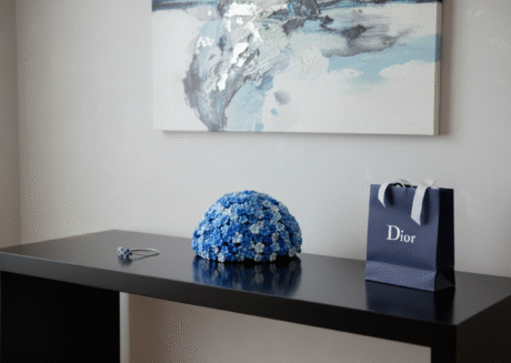 A modern room with a black console table against a light grey wall with abstract blue and silver artwork hanging above it. On the table, a unique blue floral sculpture sits prominently in the center. To its left, a delicate silver bracelet with blue accents is laid out. To the right of the sculpture, a standing blue Dior gift bag with white handles adds a touch of luxury.