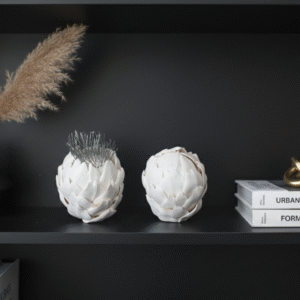 Two white ceramic artichoke sculptures, one with metal spikes emerging from the top, are displayed on a black shelf. To their left is a small black vase with a dried plant stalk, and to their right are a stack of books titled "URBAN MINIMALISM" and "FORM & LIGHT," topped with a golden abstract sculpture. The overall setting is a close-up of a minimalist designer bookcase.