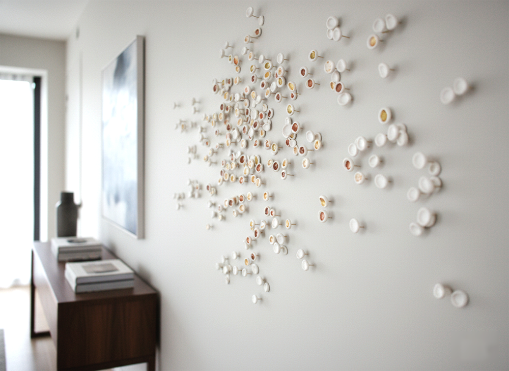 A sideways, close-up shot of a modern, minimalist living space, showcasing a unique clay wall art installation. The wall art is composed of numerous small, circular clay pieces, varying in size between 1 cm and 3 cm, attached to the light gray wall. Many of these pieces are a clean white, while others feature subtle, irregular patches of brown and yellow, suggesting a natural, earthy aesthetic. The pieces are arranged in a dynamic, organic cluster, creating a sense of movement and visual interest. To the left of the wall art, a dark wooden console table sits against the wall, holding a few decorative books and a dark, sculptural vase. Further left, a large abstract painting with cool blue and white tones hangs above the console, complementing the subtle colors of the clay art. In the background, a large window allows soft natural light to illuminate the room, highlighting the textures and forms of the artwork. The overall impression is one of sophisticated simplicity and artistic refinement.