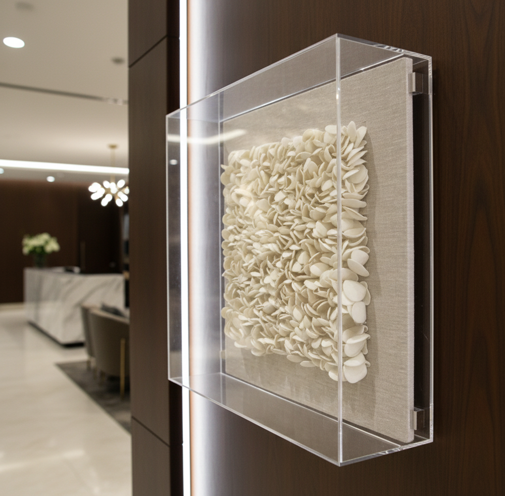 A piece of textured, off-white artwork, made of many small, overlapping petal-like shapes, is displayed within a clear acrylic (perspex) box. The box is mounted on a dark wooden wall in what appears to be a luxurious hotel reception area. The shot is a close-up, angled view that highlights the three-dimensional quality of the artwork and the reflective surface of the clear box. In the softly blurred background, elements of the hotel lobby are visible, including a reception desk and seating.