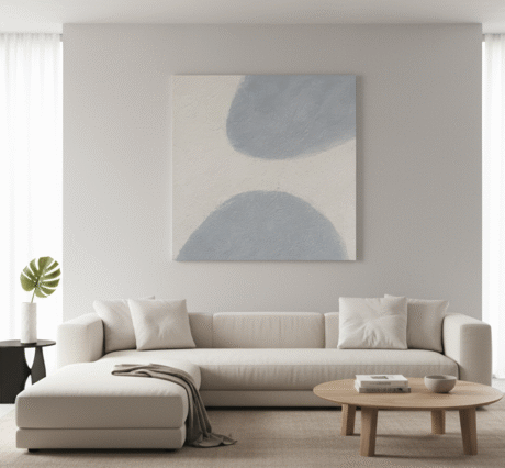 An 80x80cm painting featuring abstract blue and white shapes hangs on a light gray wall above a minimalist, off-white sectional sofa in a luxury living room. The sofa is adorned with matching pillows and a draped throw. In front of the sofa is a round wooden coffee table with books and a decorative bowl. To the left, a small black side table holds a white vase with a single monstera leaf. The room is brightly lit, with sheer white curtains on either side of the wall and a light-colored rug grounding the space.