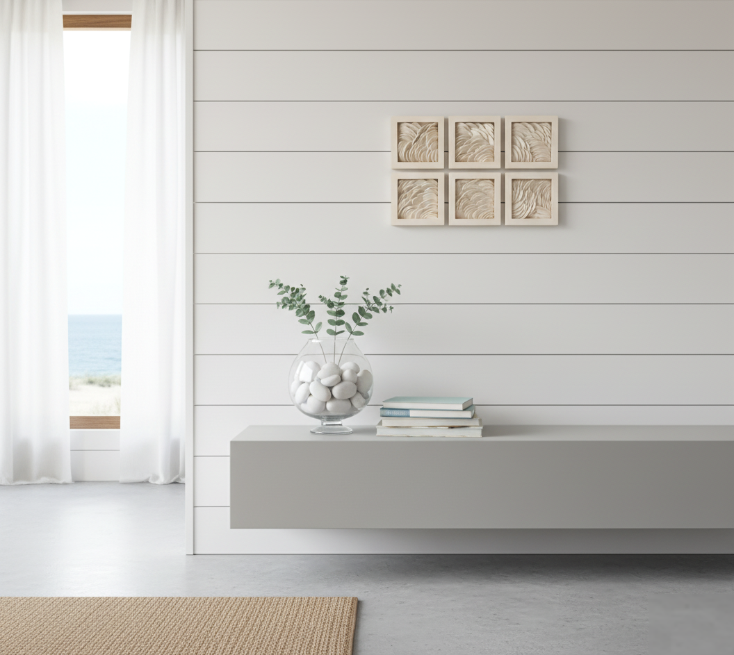 Six small, square clay artworks, each featuring a textured, wave-like pattern in off-white, are arranged in two rows of three on a shiplap wall painted in a soft, light beige. Below the artworks, a minimalist grey floating console table holds a clear glass vase filled with white pebbles and a few sprigs of eucalyptus, next to a small stack of light-colored books. To the left, a tall window with sheer white curtains offers a bright view of the ocean. The floor is a light grey concrete, partially covered by a natural fiber rug in a warm tan color.