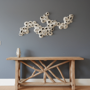 A stylish entryway features a rustic driftwood console table topped with a sleek silver vase. Above the table, a unique white coral-like sculpture, made of individual pieces arranged in an organic, flowing pattern, adorns the slate grey wall. The overall aesthetic is natural and contemporary, with warm wooden flooring extending into the background where a mirror reflects a glimpse of another room.