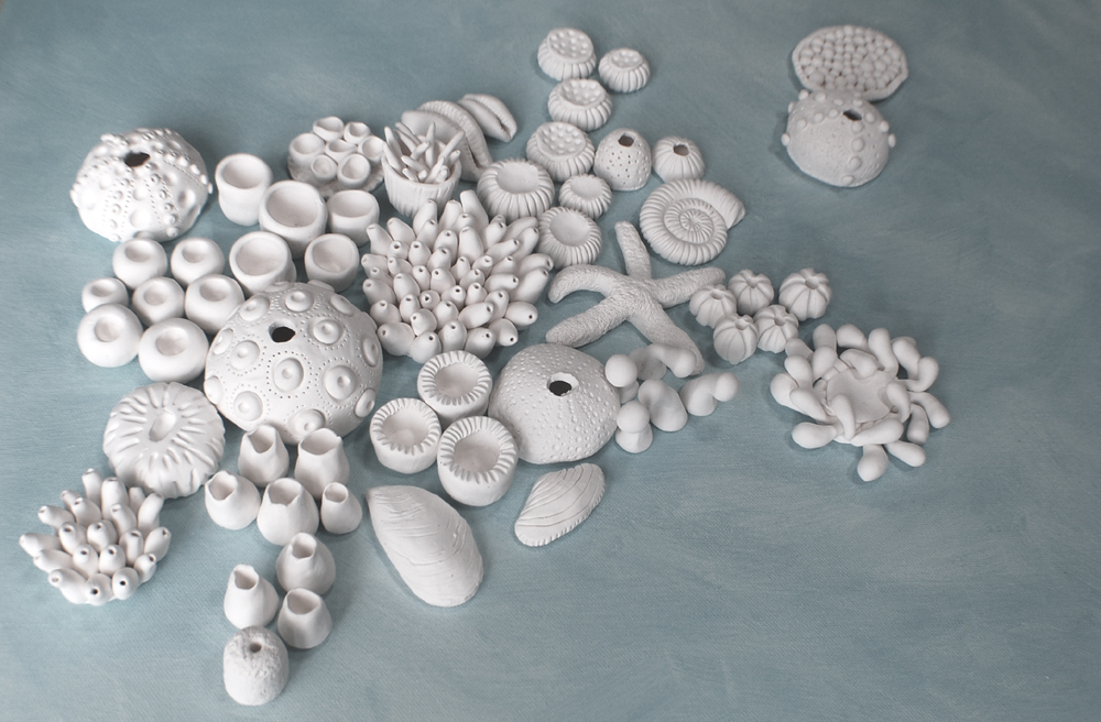 A collection of approximately 60 white, handcrafted clay marine life elements—including various coral forms, sea urchin shells, a starfish, and abstract shell shapes—artfully clustered on a pale blue textured surface, designed for a DIY wall art installation.