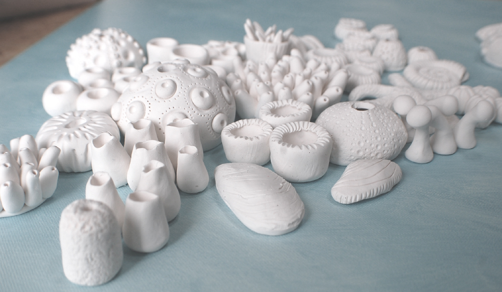A collection of approximately 60 white, handcrafted clay marine life elements—including various coral forms, sea urchin shells, a starfish, and abstract shell shapes—artfully clustered on a pale blue textured surface, designed for a DIY wall art installation.
