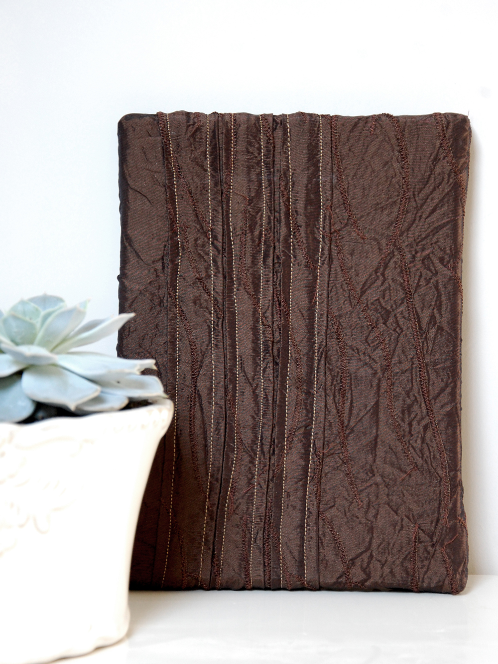 Dark brown textured taffeta wall art with vertical hand-stitching, displayed upright on a white shelf next to a potted succulent plant.
