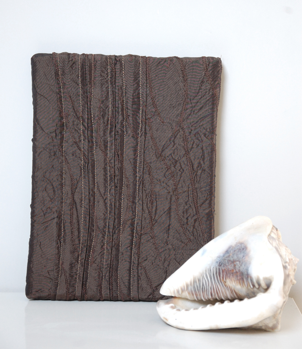 Dark brown textured taffeta wall art with vertical hand-stitching, displayed upright on a white shelf next to a potted succulent plant.
