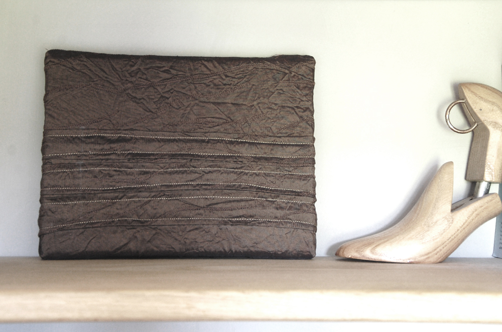 Dark brown textured taffeta wall art with vertical hand-stitching, displayed upright on a white shelf