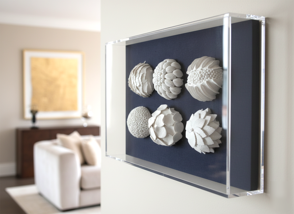 Six white, textured, coral reef-inspired sculptures are displayed within a clear acrylic box frame against a dark blue background. The frame is mounted on a light-colored wall, with a soft-focused luxury interior visible in the background, including a cream-colored sofa, a dark wooden console table, and a gold-framed artwork.