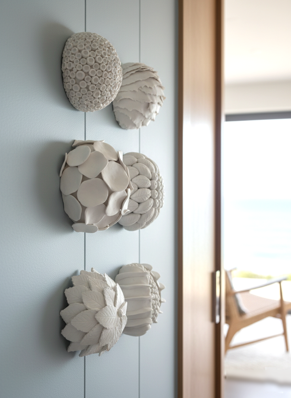 Six white coral reef-inspired wall sculptures are arranged vertically on a light blue wall in a luxury coastal interior. The sculptures are textured with organic patterns, resembling various marine forms. In a sideways close-up shot, the topmost sculpture has a bumpy, brain-like texture, while the one below it features ruffled, layered edges. The middle two sculptures are composed of overlapping circular and leaf-like shapes, creating a sense of depth. The bottom two sculptures exhibit more defined leaf and ridged patterns. The overall arrangement measures approximately 14 cm in width, with each sculpture being about 6 cm in size. The background is softly blurred, revealing a wooden door frame, a glimpse of a modern chair, and a bright window looking out onto the ocean, emphasizing the coastal setting. The natural light highlights the intricate details and delicate shadows of the sculptures.