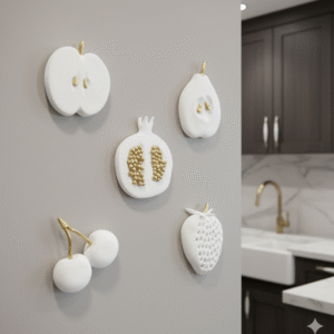 Five white fruit sculptures with gold accents, including an apple, pear, pomegranate, cherries, and a strawberry, are arranged on a light gray painted wall in a luxurious kitchen. The sculptures, each measuring about 8cm, are captured in a close-up shot, with a glimpse of dark wood cabinets, a white sink, and a gold faucet in the background, maintaining a realistic scale.