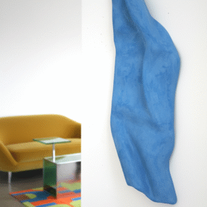 A vertical blue sculpture with a textured surface and organic, wave-like contours is mounted on a stark white wall. The sculpture is predominantly in focus, highlighting its three-dimensional form and rough, matte finish. In the blurred background to the left, a modern room is visible with a mustard yellow sofa, a reflective side table, and a colorful rug with geometric patterns in shades of orange, green, and blue. The overall impression is an artistic piece showcased in a contemporary, stylish setting.