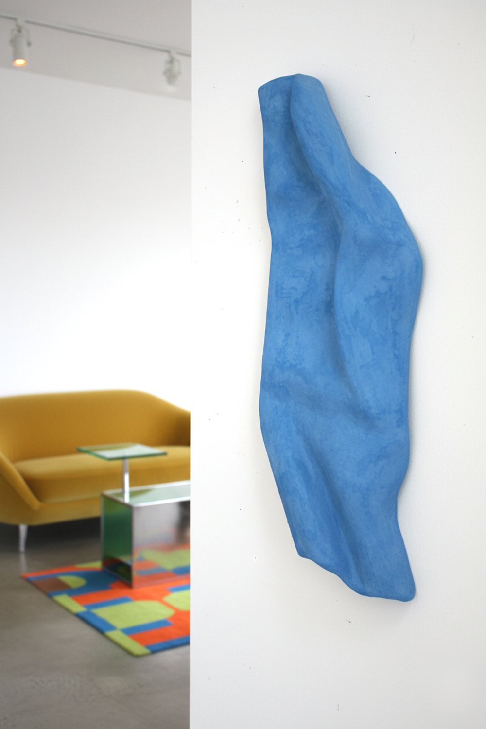 A vertical blue sculpture with a textured surface and organic, wave-like contours is mounted on a stark white wall. The sculpture is predominantly in focus, highlighting its three-dimensional form and rough, matte finish. In the blurred background to the left, a modern room is visible with a mustard yellow sofa, a reflective side table, and a colorful rug with geometric patterns in shades of orange, green, and blue. The overall impression is an artistic piece showcased in a contemporary, stylish setting.