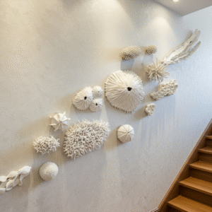 The image shows an intricate clay wall installation containing 14 white intricate sculptures depicting coral reef textures or elements. They have been hung on a staircase on the left. On the right is a wooden staircase. Overall feel is luxurious and high-end.