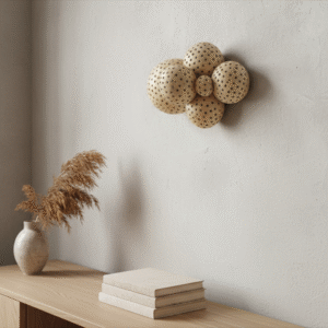 A Japandi-style living room features an abstract artwork composed of light brown, perforated spheres arranged in a cluster on a textured beige wall. Below the artwork, a light wood console table holds a stack of three neutral-toned books and a cream-colored vase with dried pampas grass. The room has soft, diffused lighting, creating a serene and minimalist atmosphere.