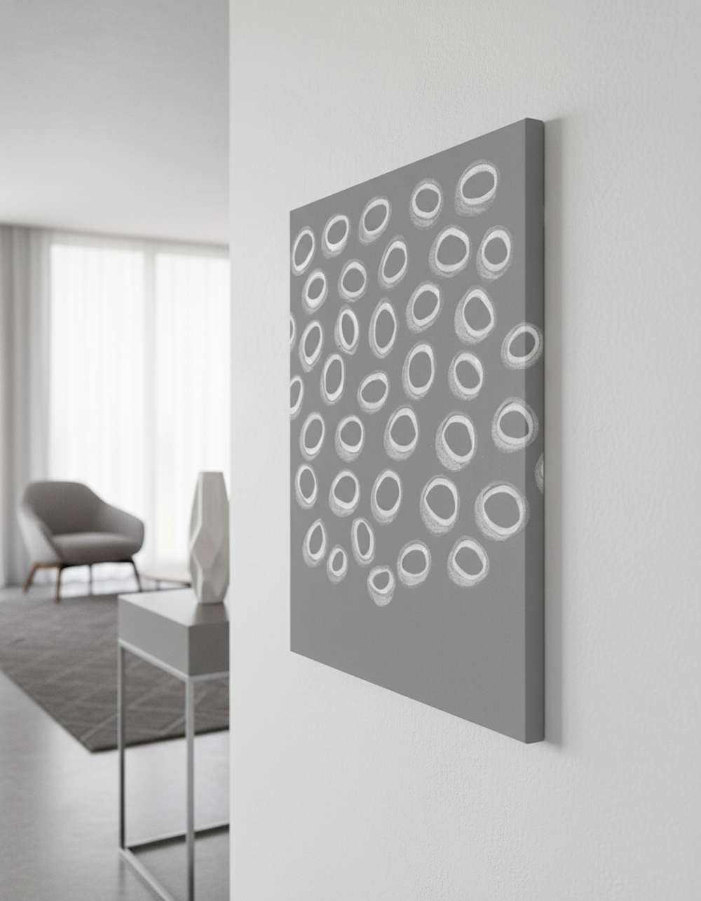 A close-up, sideways shot shows a 70 x 50 cm gallery-wrapped canvas hanging on a white wall in a luxurious, white and gray interior. The canvas features a painting of various white, hand-drawn oval shapes on a gray background, suggesting an abstract and organic pattern. In the softly blurred background, a contemporary gray armchair, a light-colored sculptural vase on a minimalist side table, and a large window with sheer white curtains are visible, enhancing the sophisticated and elegant atmosphere of the space.
