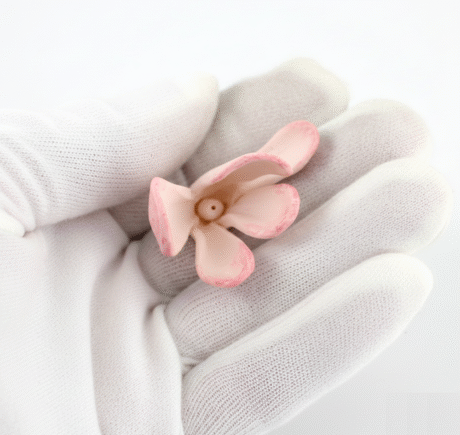 A close-up shot features a delicate, light pink floral sculpture, possibly made of porcelain or ceramic, resting gently in the palm of a hand encased in a pristine white glove. The hand is angled slightly towards the camera, showcasing the intricate details and soft hues of the sculpture against the clean, white fabric of the glove. The lighting is even, highlighting the textures and contours of both the sculpture and the glove.